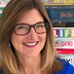 Smiling woman with glasses and straight, shoulder-length hair, standing in front of shelves filled with board games.