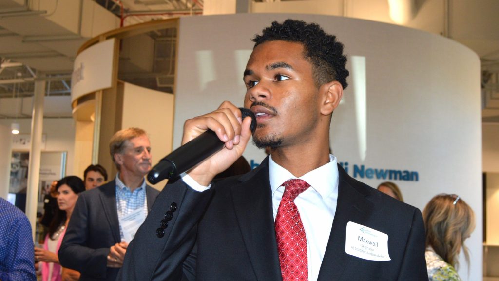 A person in a suit holds a microphone, speaking at an event with attendees in the background.