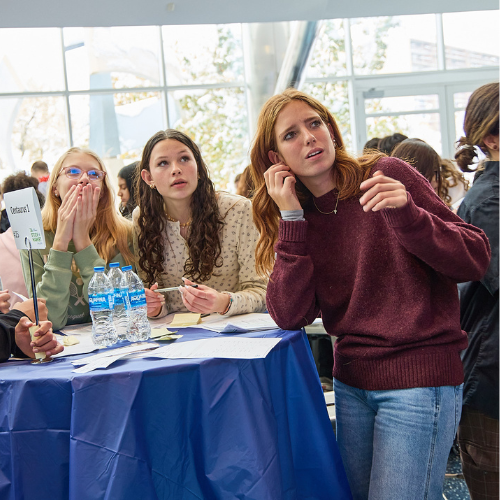 People at a table appear engaged, holding notes and looking towards an event, with water bottles and papers on the table.