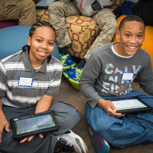 Two kids sitting and smiling while using tablets, wearing casual clothes and name tags, with others seated nearby.