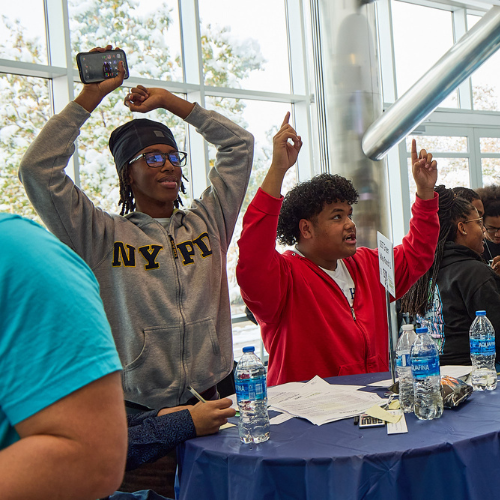 Two people joyfully raise their arms while seated at a table with water bottles inside a sunlit room.